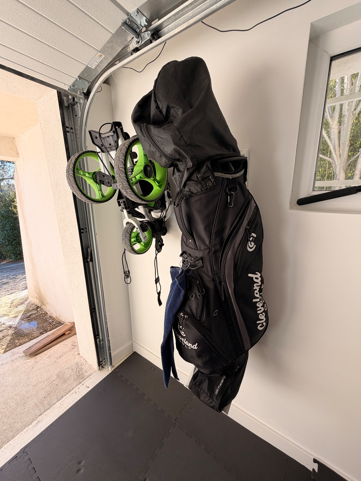 Sac de golf noir suspendu à un mur, avec un support de caddy et un coin de garage visible.